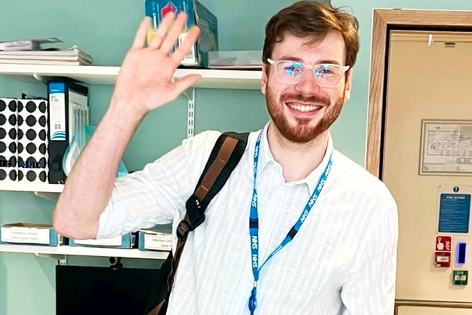 A man smiling, wearing a light blue shirt, waves at the camera, standing inside an office workspace.
