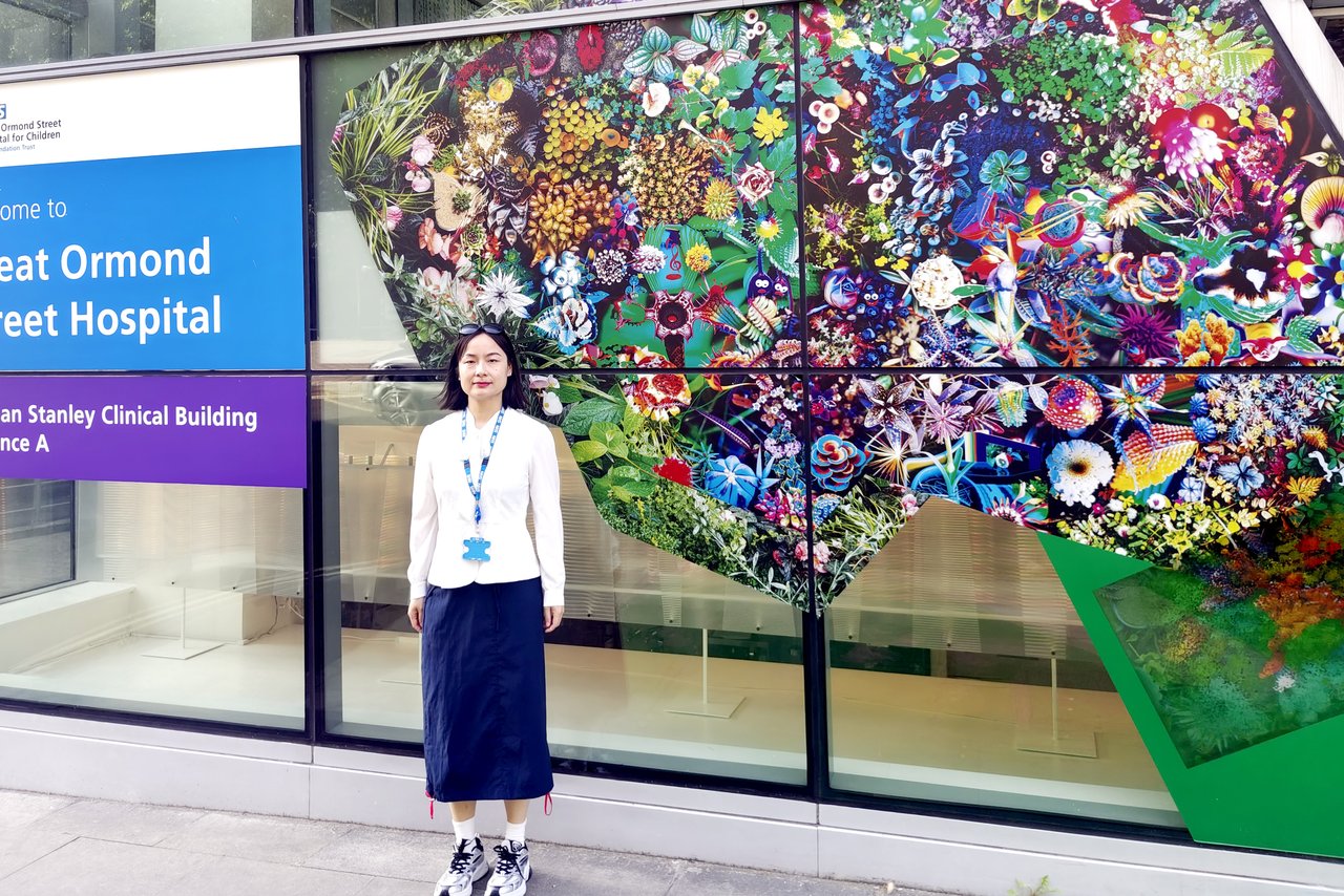A woman wearing a white top and dark skirt stands outside Great Ormond Street Hospital, beside the Morgan Stanley Clinical Building entrance, in front of a large colourful nature-themed artwork displayed on the glass façade.