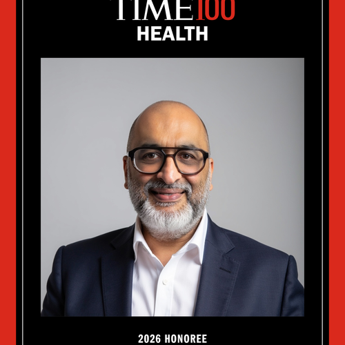 A black and red framed head shot of a man with thick black glasses smiling at the camera
