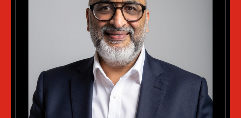 A black and red framed head shot of a man with thick black glasses smiling at the camera
