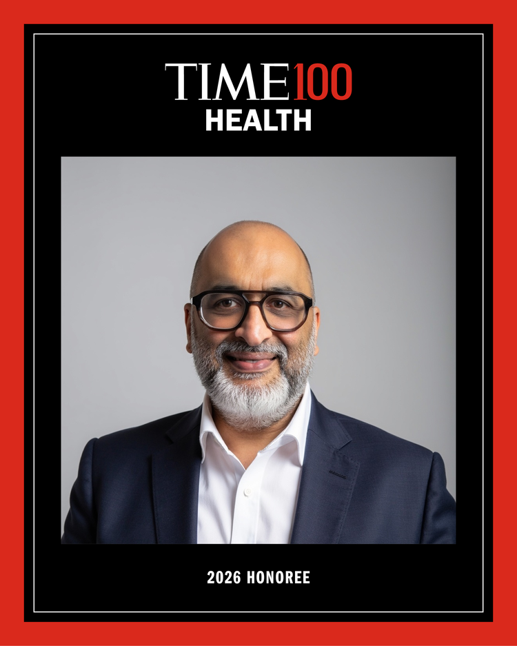 A black and red framed head shot of a man with thick black glasses smiling at the camera