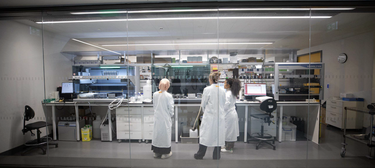 Three people in white lab coats stand in a modern laboratory, viewed through a glass wall, facing a long bench lined with automated equipment, monitors, bottles, and pipetting machines.
