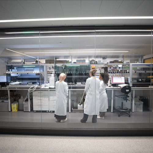 Three people in white lab coats stand in a modern laboratory, viewed through a glass wall, facing a long bench lined with automated equipment, monitors, bottles, and pipetting machines.