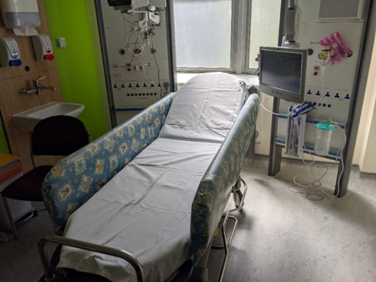 A hospital room with a single bed covered in a blue and white patterned blanket, positioned near a window. To the right of the bed is medical equipment including a monitor and tubes. On the left side, there is a sink with wall-mounted soap dispensers and an office chair. The walls are painted green and white.