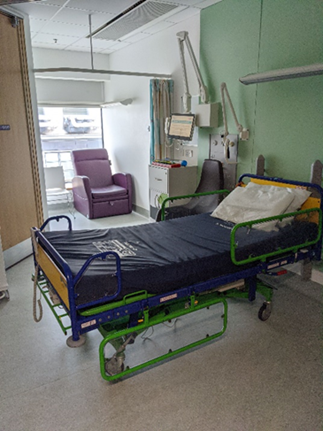 Hospital room with a bed featuring green and blue railings and a pillow. In the background, a purple chair sits beside a window. Medical equipment, including monitors and cables, is mounted on the wall.
