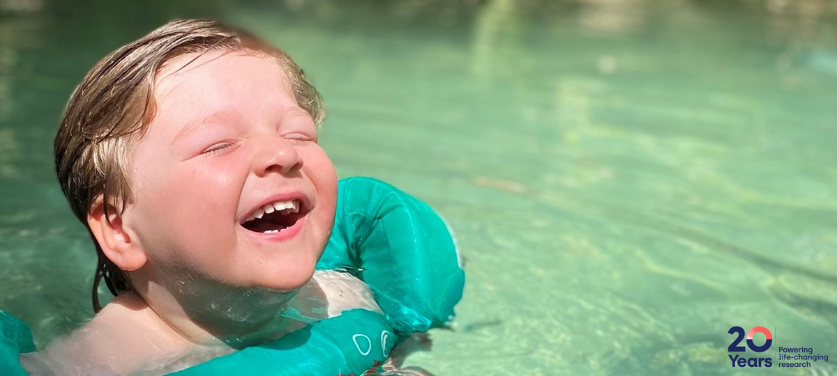 A young boy swimming. He is wearing green arm bands and smiling. His eyes are closed.