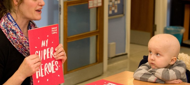 A women in a black cardigan and flower top showing a pink workbook saying 'My Super Heroes' to a child with a grey jumper on.