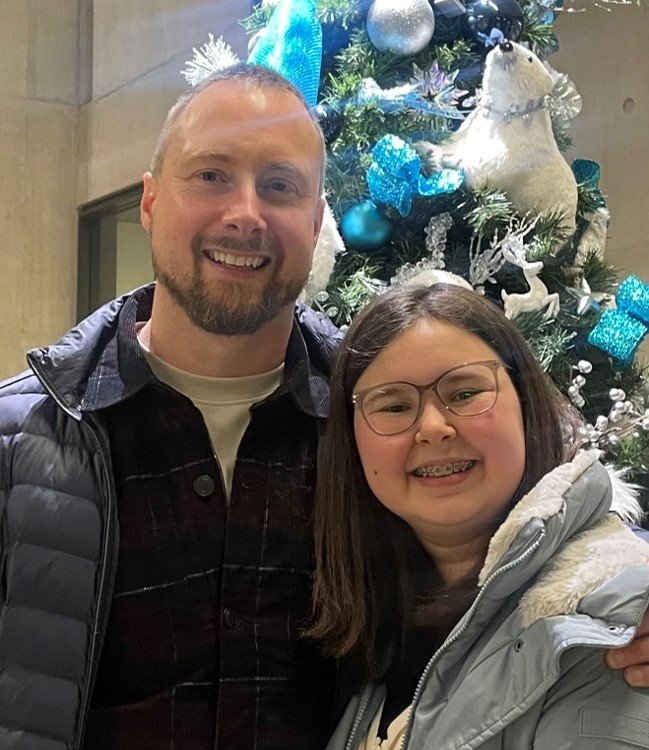 A father and daughter are wearing coats for winter and smiling broadly in front of a Christmas tree decorated in blue and silver