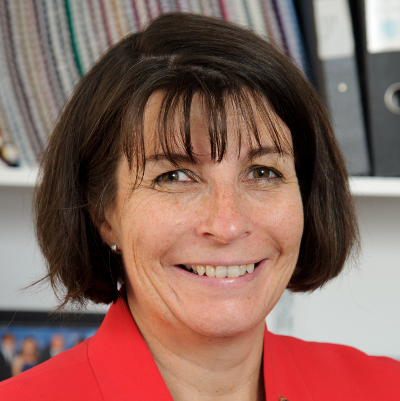 A woman is stood smiling at the camera in a red jacket and shelves of dossiers behind her.