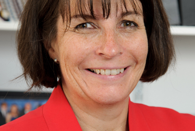 A woman is stood smiling at the camera in a red jacket and shelves of dossiers behind her.