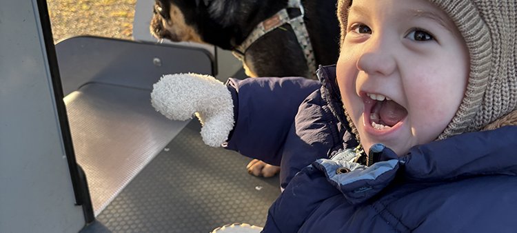 A little boy in hat and big coat has his mouth wide open, maybe squealing, at the camera and you can see his black small dog behind him