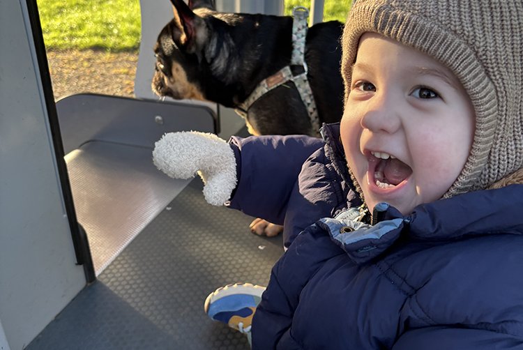 A little boy in hat and big coat has his mouth wide open, maybe squealing, at the camera and you can see his black small dog behind him