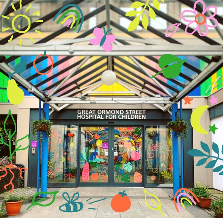 Entrance of Great Ormond Street Hospital for Children, featuring vibrant and whimsical decorations such as suns, rainbows, butterflies, flowers, leaves, and fruits. The hospital name is prominently displayed above glass doors beneath a glass canopy supported by blue pillars, with hanging flower baskets flanking the entrance.