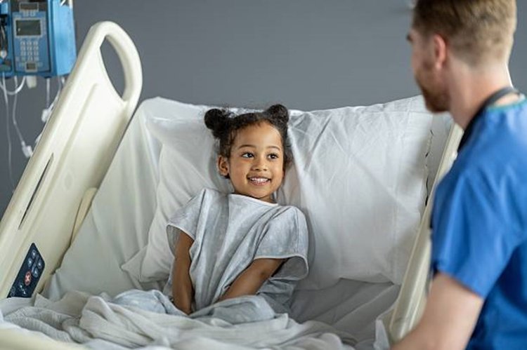 A young child lying in a hospital bed, wearing a hospital gown and covered with a blanket.