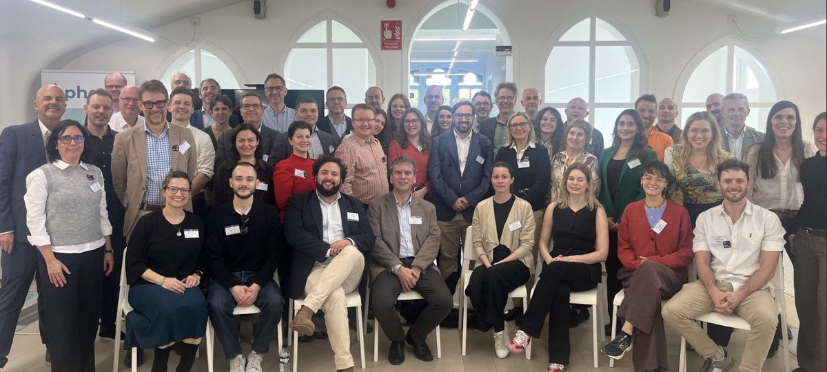 large group of roughly 50 adults gathered in a bright white room with some sat in chairs, smiling at camera.