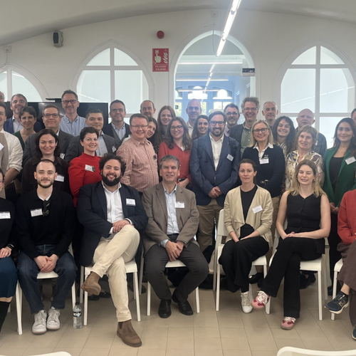 large group of roughly 50 adults gathered in a bright white room with some sat in chairs, smiling at camera.