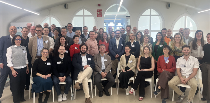 large group of roughly 50 adults gathered in a bright white room with some sat in chairs, smiling at camera.
