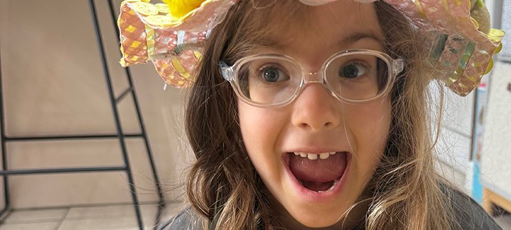 A young girl in a flowery easter bonnet smiles broadly at the camera with her mouth open wide
