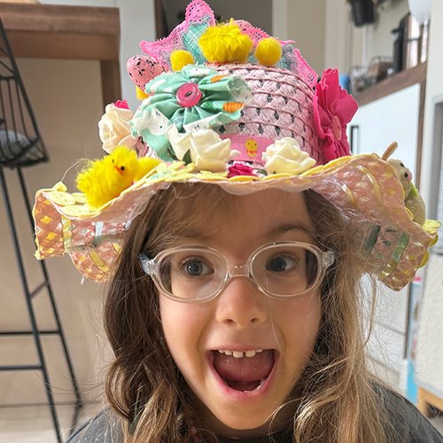A young girl in a flowery easter bonnet smiles broadly at the camera with her mouth open wide