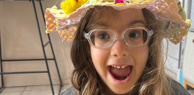 A young girl in a flowery easter bonnet smiles broadly at the camera with her mouth open wide