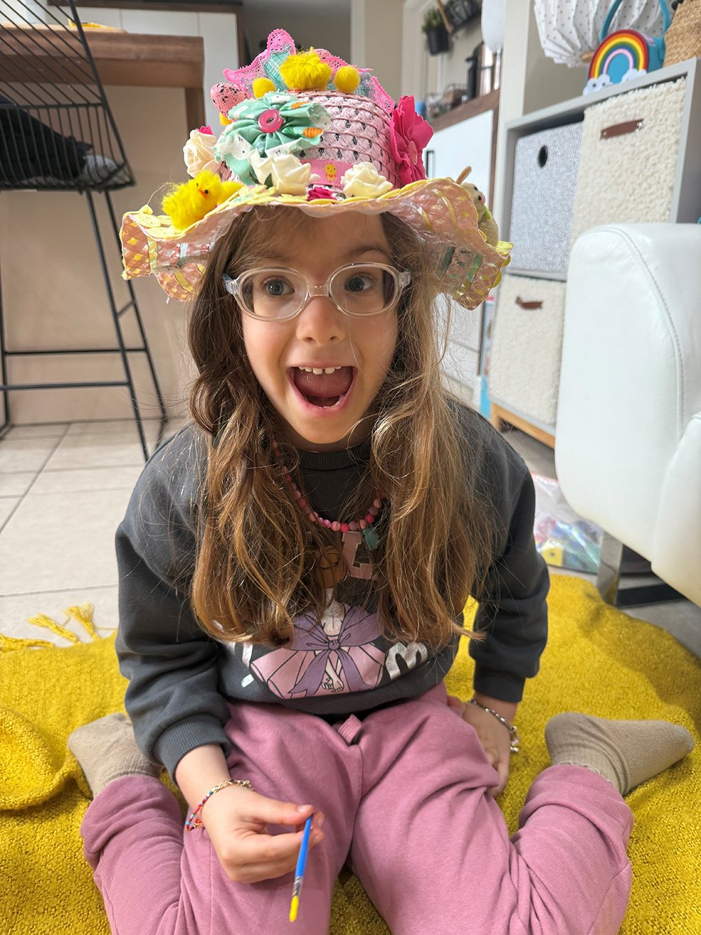 A young girl in a flowery easter bonnet smiles broadly at the camera with her mouth open wide