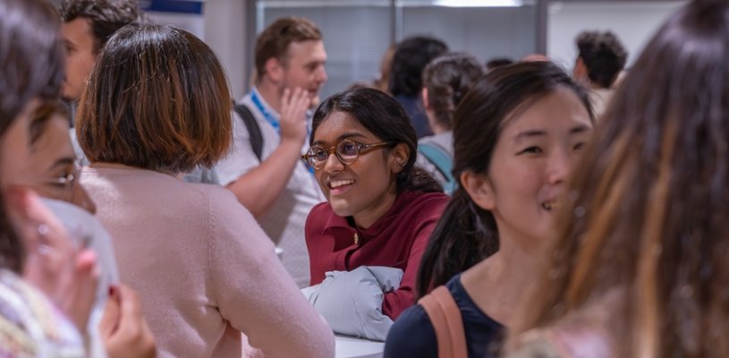 A Crowd of people talk in a busy room - the central focus shows a woman smiling and talking with a colleague who has their back to the camera