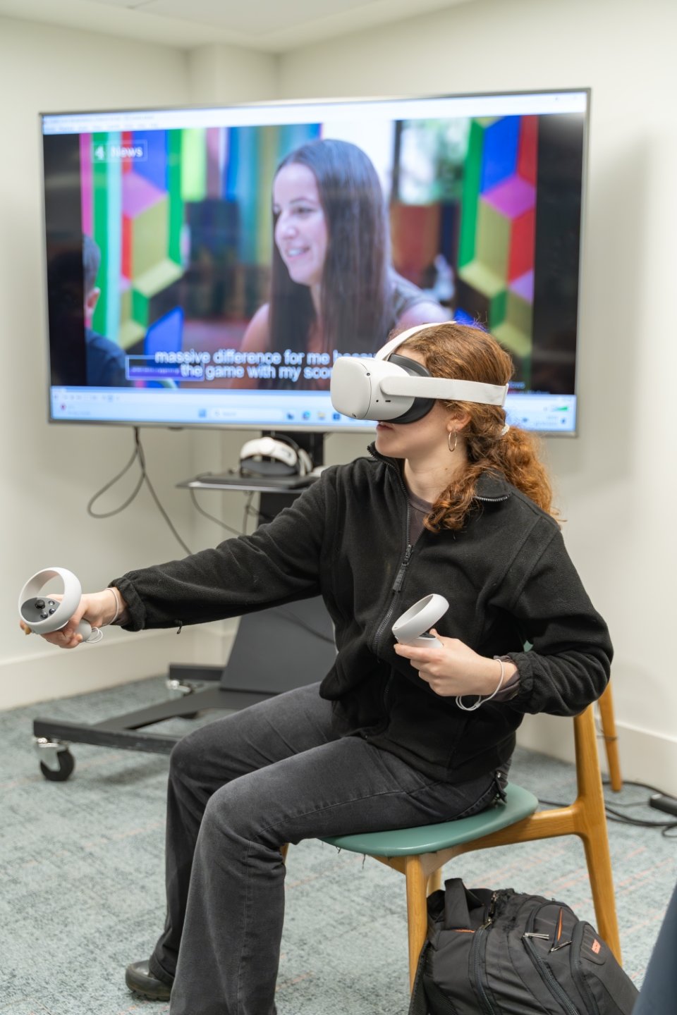 An adolescent with ginger hair tied back in a pony tail wears a virtual reality headset and holds controllers in her hands